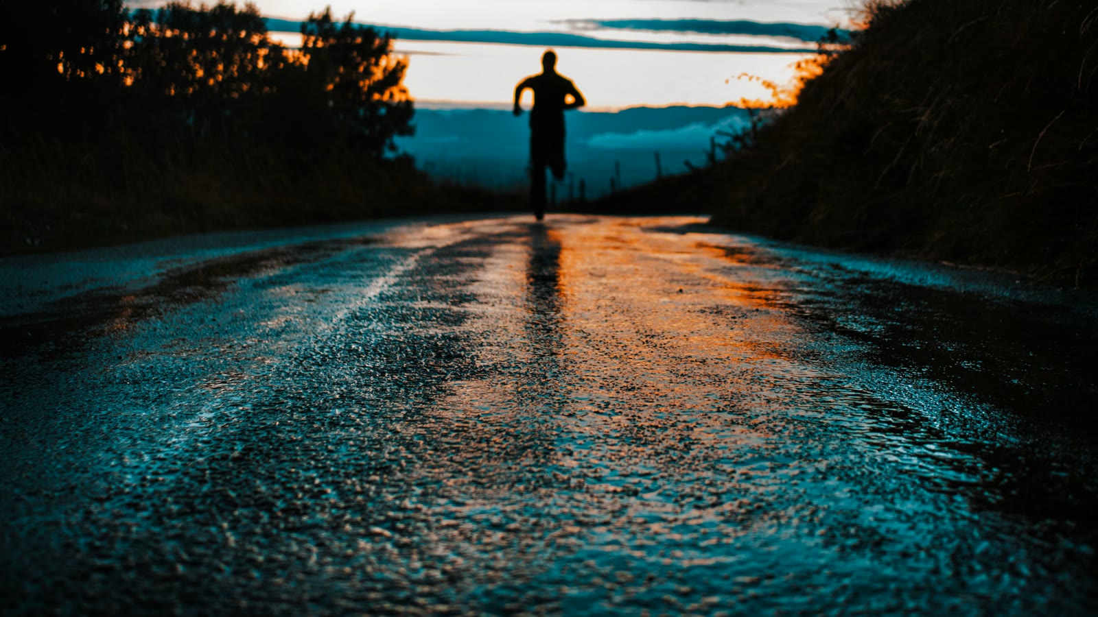A runner silhouetted on a wet road at dawn, the sky reflected in the water